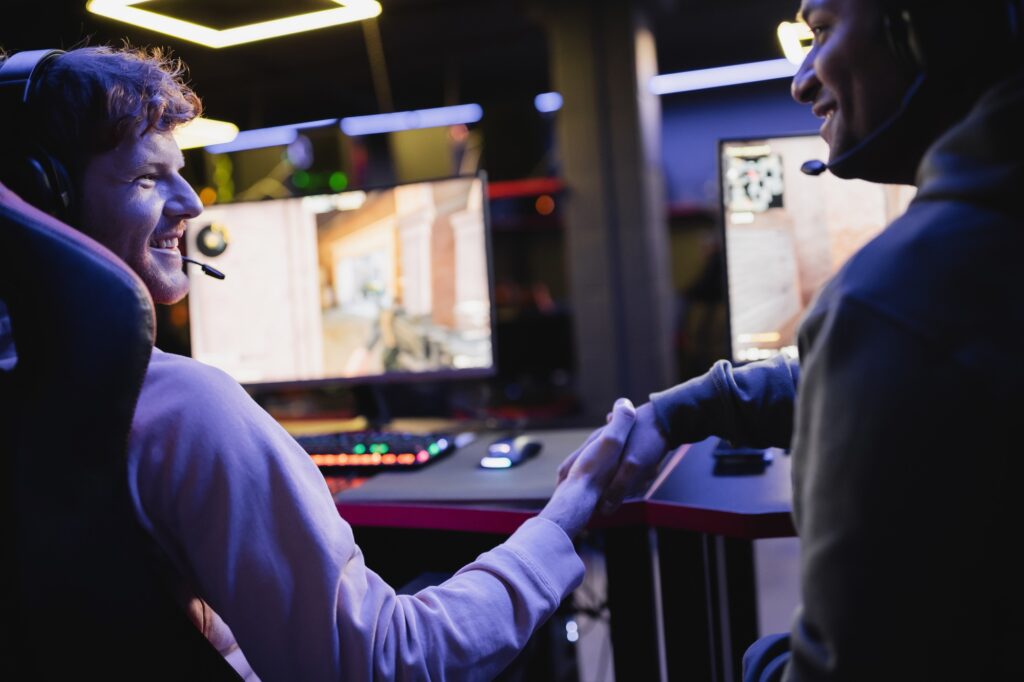 Smiling multiethnic gamers shaking hands near blurred computers in gaming club